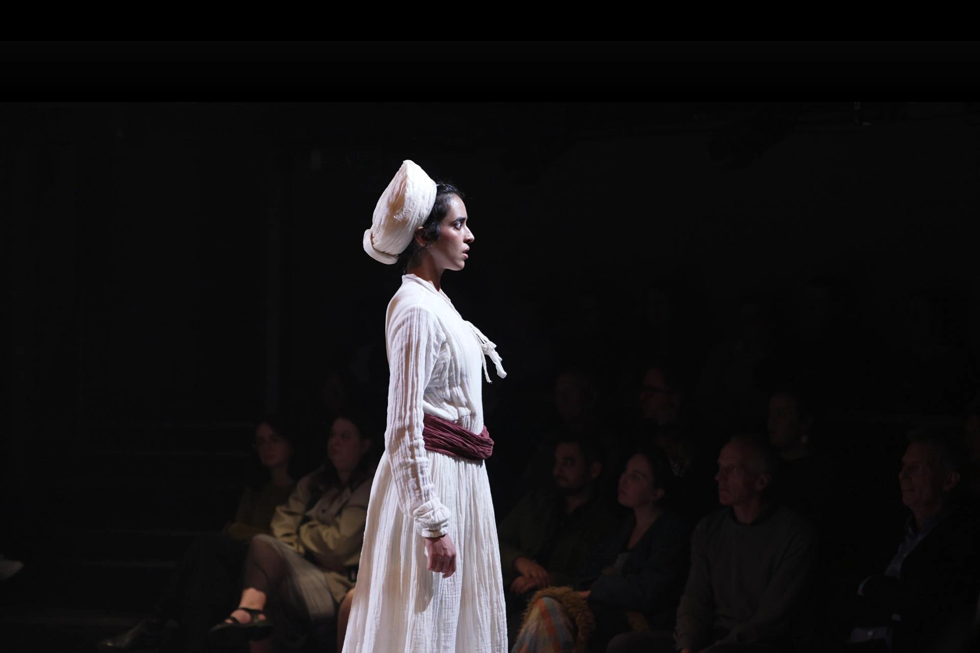 An actor in period nurse costume, spotlit on stage in front of an audience